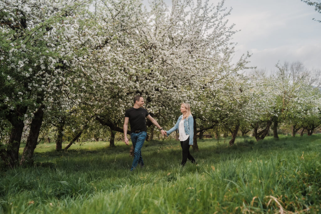 Couple walking hand in hand through blooming apple orchards during a relaxed outdoor portrait session in Switzerland.